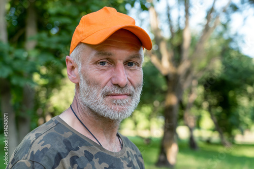 Close up portrait of sad mature man wearing orange cap looking at camera, standing in park outdoors. Senior caucasian man with grey beard.