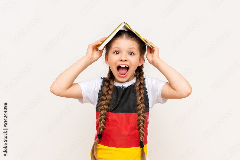 A little girl with a book on her head on a white isolated background. Learning German in the lower grades. A child in a T-shirt with the flag of Germany. A little girl with a book on her head on a white isolated background. Learning German in the lower grades. A child in a T-shirt with the flag of Germany.