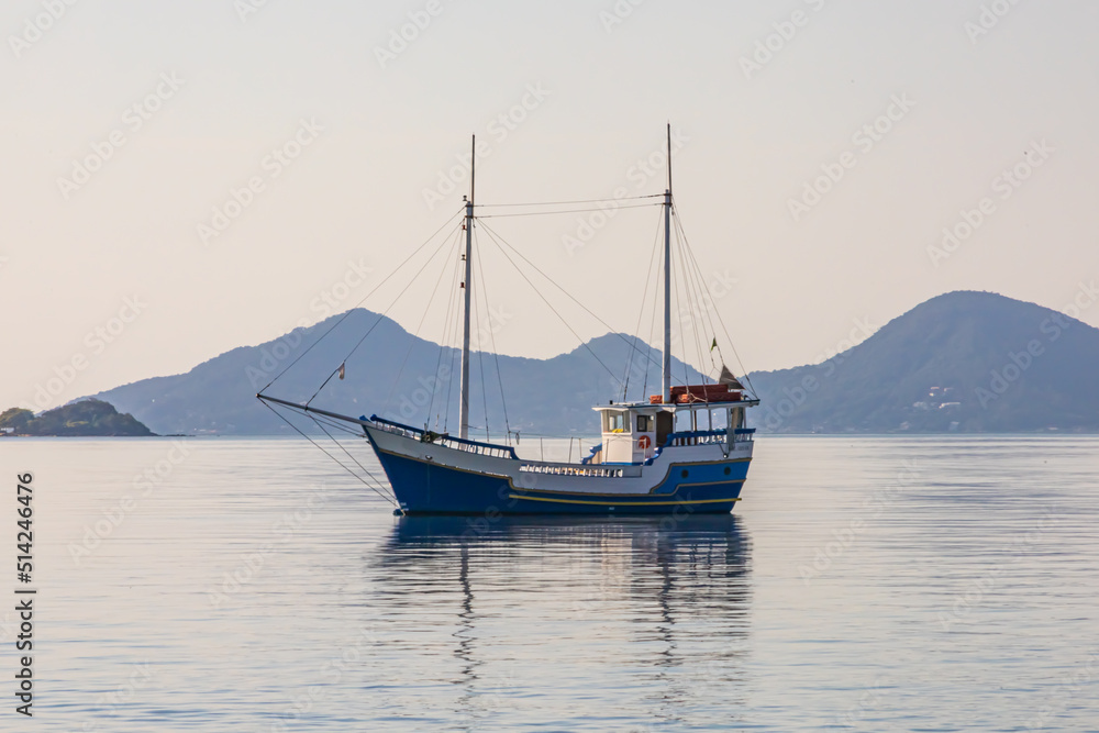 Sailing ship on the smooth water of the sea. Windless sea. There are no waves. Hills and mountains on the horizon. Blue sky. A ship with lowered sails is at anchor. Reflection of the ship on the water