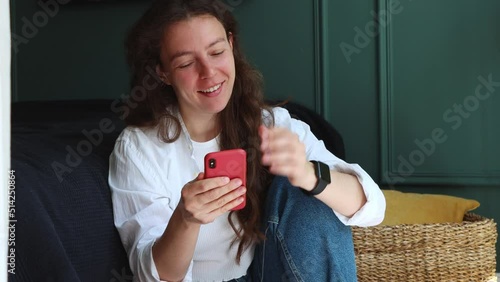 Happy pretty brunette girl using mobile phone, virtual chatting with boyfriend, sitting indoors in green room, wearing white shirt. Feeling enjoy, reading good news in message, online browsing app. photo