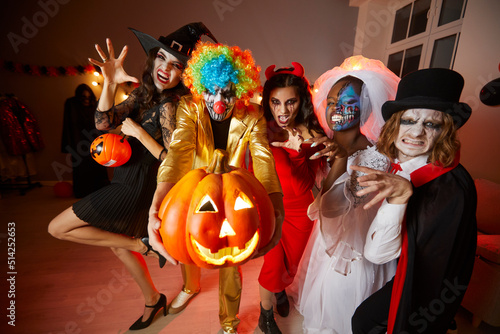 Group of young happy friends in costumes on Halloween having fun at party making scary faces. Cheerful multiethnic people with glowing jack-o-lantern doing claw hand gesture looking at camera.