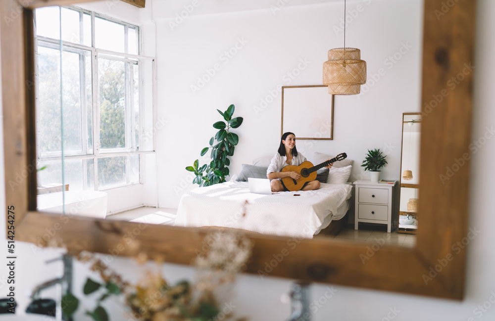 Fototapeta premium Brunette female learning how to play guitar while watching online video course on laptop computer in cozy home interior. Young woman with netbook enjoying hobby with musical instrument relaxing on bed