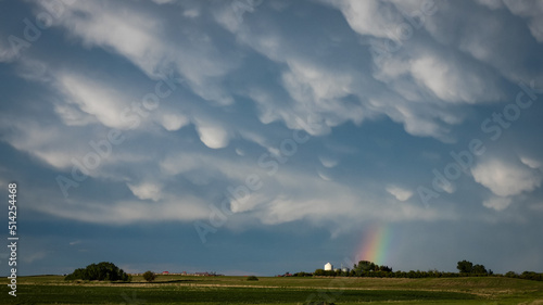 Storm clouds above farmland with a small rainbow peaking out below the fluffy Mammatus clouds
