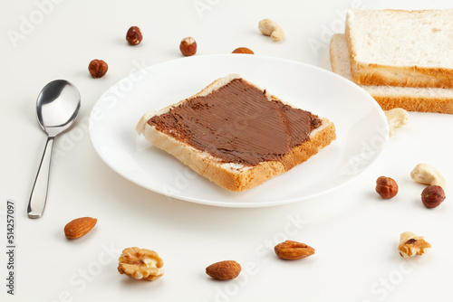 Square bread for toast with chocolate spread on a plate. Nuts, a spoon, slices of bread and a white plate with a sandwich on a white table.
