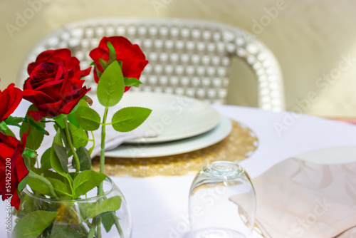 Date table. Beautiful table setting at an expensive restaurant. Red roses in a vase decorates the tablecloth. Plates, glasses stand on the table. Wedding celebration. Classical serving in cafe terrace