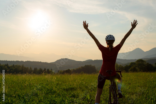Female cycle race rider pedaling on an open road route in mountain landscape, passing by a car and raising arms in a victory pose, wide shot.
