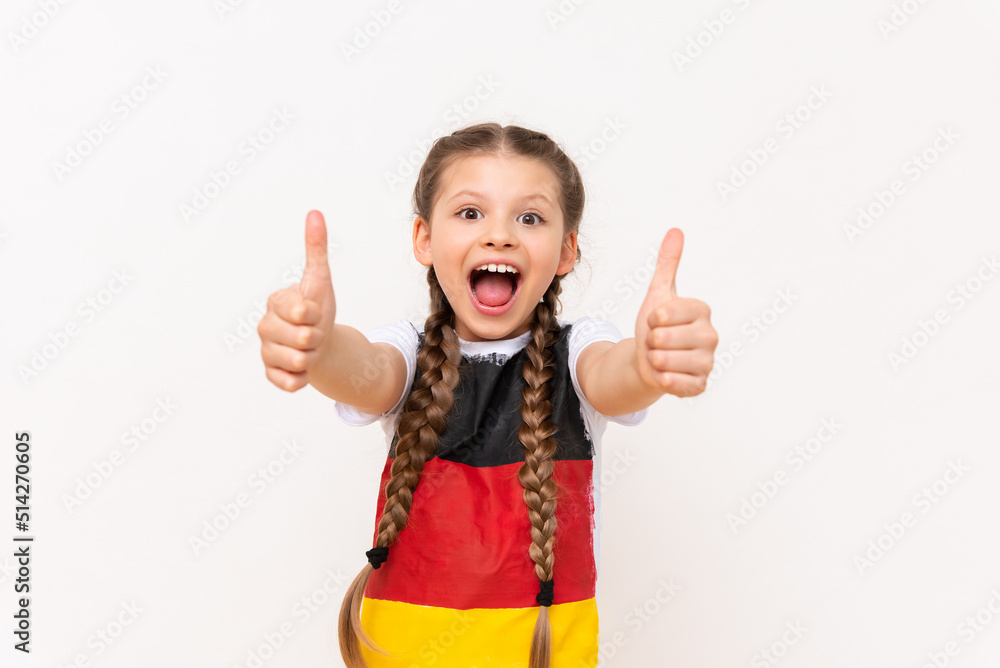 A little girl with a German flag on a T-shirt, with long hair in pigtails, gives a thumbs up and smiles broadly on a white isolated background. Language courses. A little girl with a German flag on a T-shirt, with long hair in pigtails, gives a thumbs up and smiles broadly on a white isolated background. Language courses.