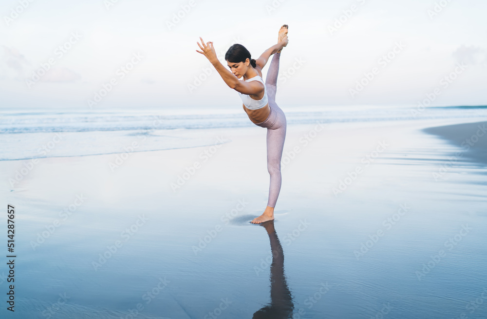 Obraz premium Fit girl in sportive tracksuit raising leg while doing balance asana for stretching and recreate, attractive woman practice harmony meditation in yoga pose spending weekend leisure for training
