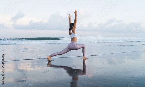 Side view of young Caucasian female yogi stretching legs and body muscles during morning training at coastline beach, flexible woman in sportive tracksuit standing in hatha asana at seashore