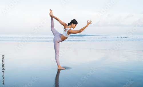 Caucaisan woman with good body shape concentrated on hatha asana pose with raised leg, young female in sportive tracksuit stretching for feeling vitality during holistic healing on seashore retreat