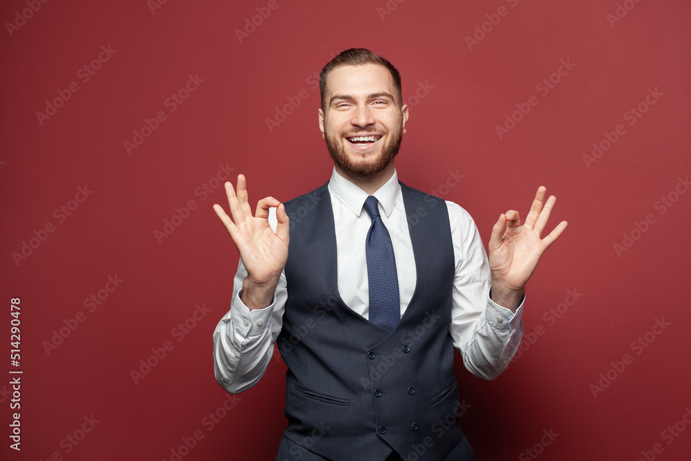 Portrait of a smiling businessman dressed in suit showing ok gesture on red background Portrait of a smiling businessman dressed in suit showing ok gesture on red background
