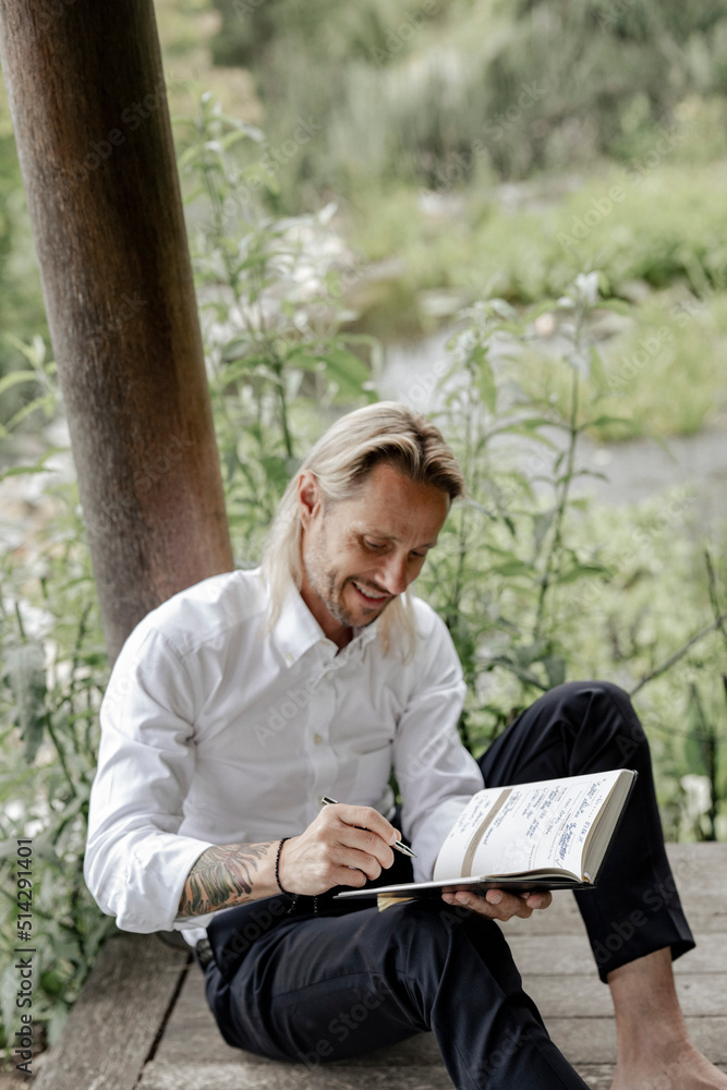 Geschäftsmann im weißen Hemd sitzt auf Holzsteg und schreibt in ein Buch