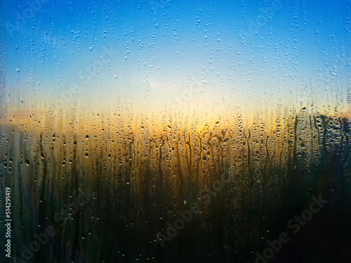 Drops flowing from fogged glass and behind glass rising sun with sky in blue and yellow tones. Texture of dripping drops on damp glass.