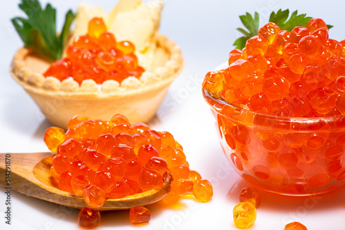 Delicious red caviar in wooden brown spoon, in small crystal cup with top decorated with parsley and red caviar in tartlet with butter and parsley on white background.