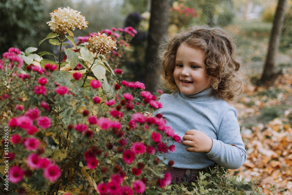 adorable curly little girl in blue shirt in park with flowers at fall time, autumn, health, card, banner