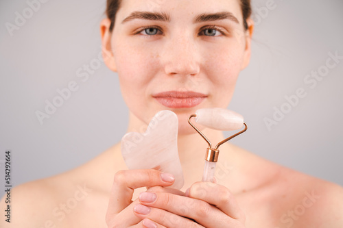 Caucasian woman holding pink roller massager and gouache scraper on white background. 