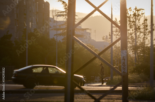 Sunset in a city with trees and residential buildings, while a young man waits standing in front of a car