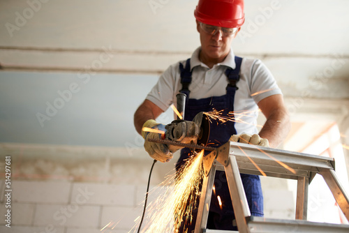 Fototapeta Professional construction worker hands in work gloves using angle grinder to cut metal rod at building site