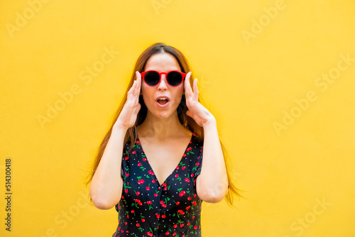 portrait of a young brunette woman isolated on yellow background looking surprised. happy girl wearing summer clothes and sunglasses in a good mood and smiling. joy, fun, youth and lifestyle concept