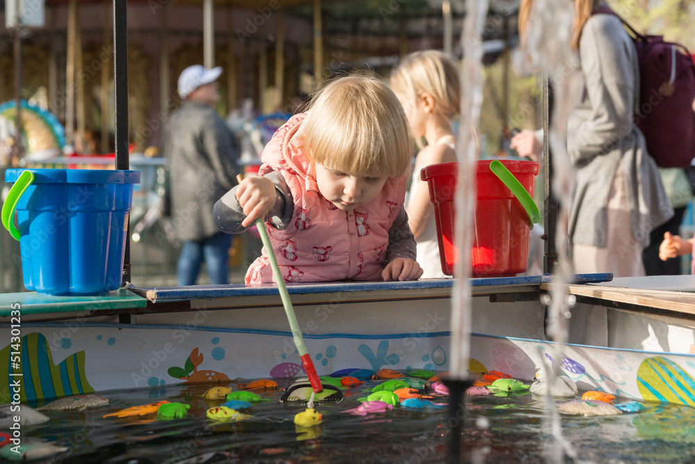 Fototapeta premium A serious girl in a city park is playing fishing with magnetic fishing rod and toys