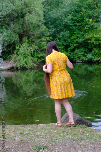 Fototapeta Naklejka Na Ścianę i Meble -  happy young woman with long hair in a yellow dress stands on the banks of a river, a lake and looks into the water, rear view, the concept of a happy female life, an active lifestyle, healthy hair