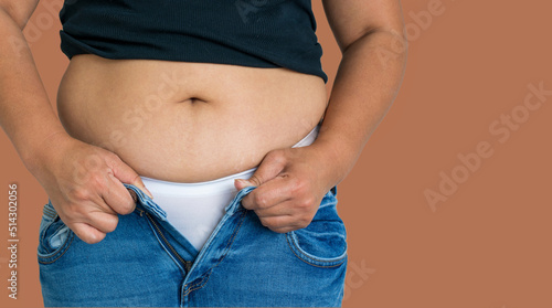 Cropped mid section of an obese overweight woman trying to squeeze her big body into small size jeans isolated on brown background, copy space. Concept for losing weight. 