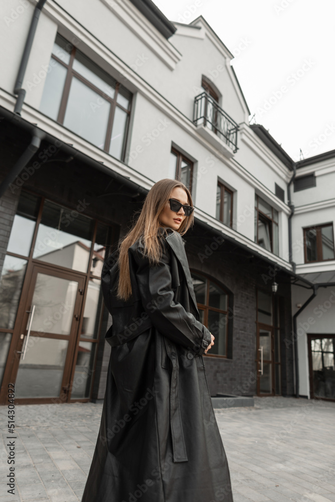 Fashionable young pretty woman with vintage sunglasses in stylish black long leather coat walks  near a building on the street