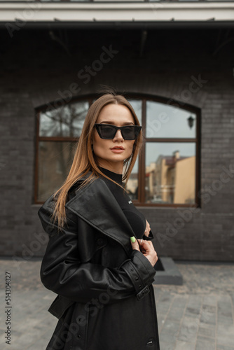 Fashionable portrait of beautiful young woman with black fashion glasses in black stylish leather coat poses in the city