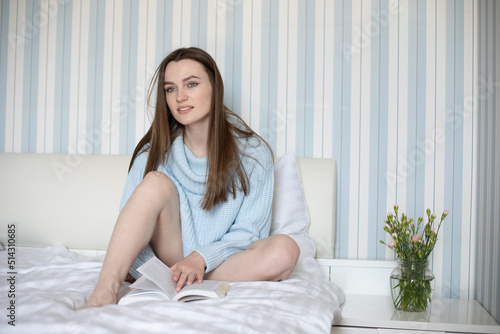 Attractive long-haired brown-haired woman in blue sweater is sitting on bed in blue and white bedroom with book.  © Татьяна Волкова