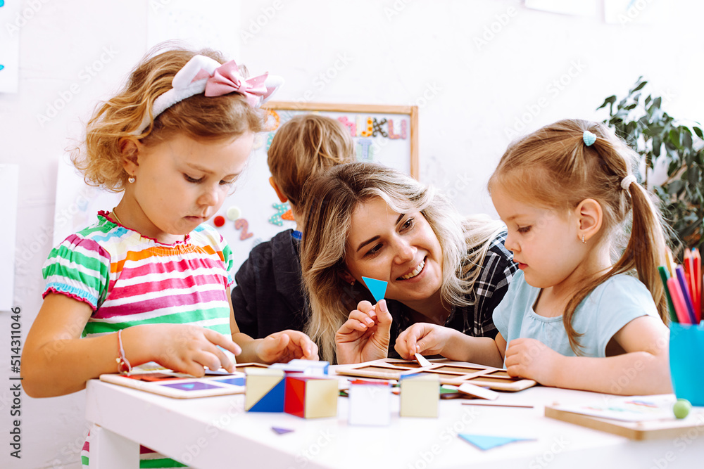 Little girls and smiling educator studying geometric figures, playing and folding cubes on desk sitting on floor in playroom. Interesting lesson for kindergartners developing logic and intelligence - obrazy, fototapety, plakaty 