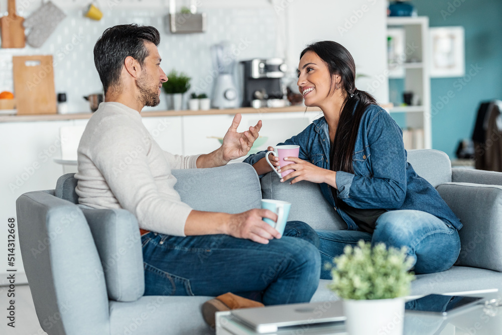 Obraz premium Lovely couple holding coffee cups while talking sitting on couch at home.