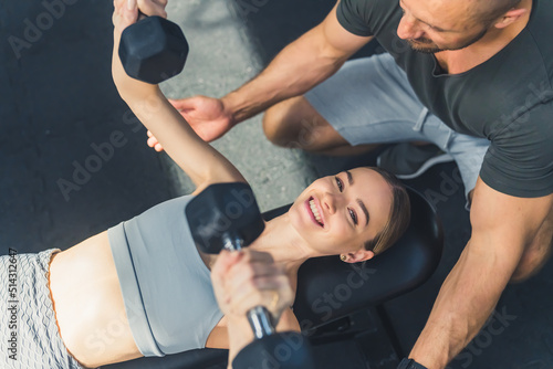 Indoor shot of a cheerful caucasian active woman in a sports outfit lifting light dumbbells lying on her back. Muscular man helping her. High quality photo