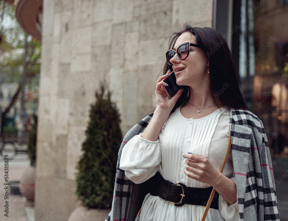 Fototapeta premium Attractive cheerful young woman in sunglasses speaking on cellphone in town street and smiling.