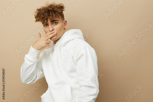 an attractive man stands on a beige background in a white hoodie and looking at the camera touches his mouth with two fingers