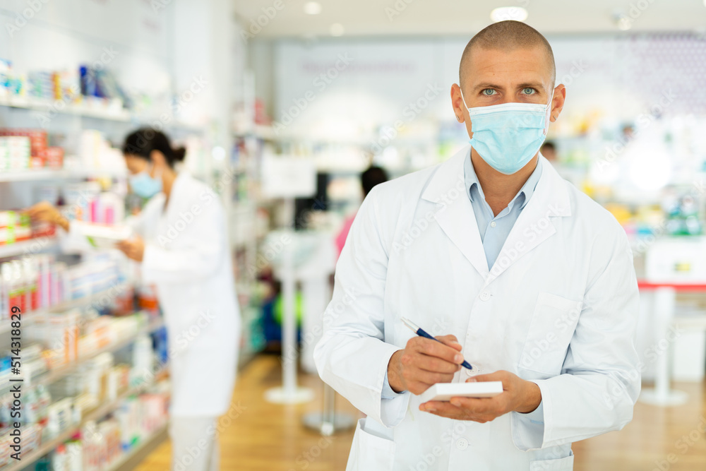 Portrait of male pharmacist in protective mask, working in pharmacy during the pandemic standing in trading floor and makes important notes. High quality photo