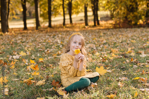 A beautiful little blonde girl in a yellow dress sits in an autumn park holding maple leaves.