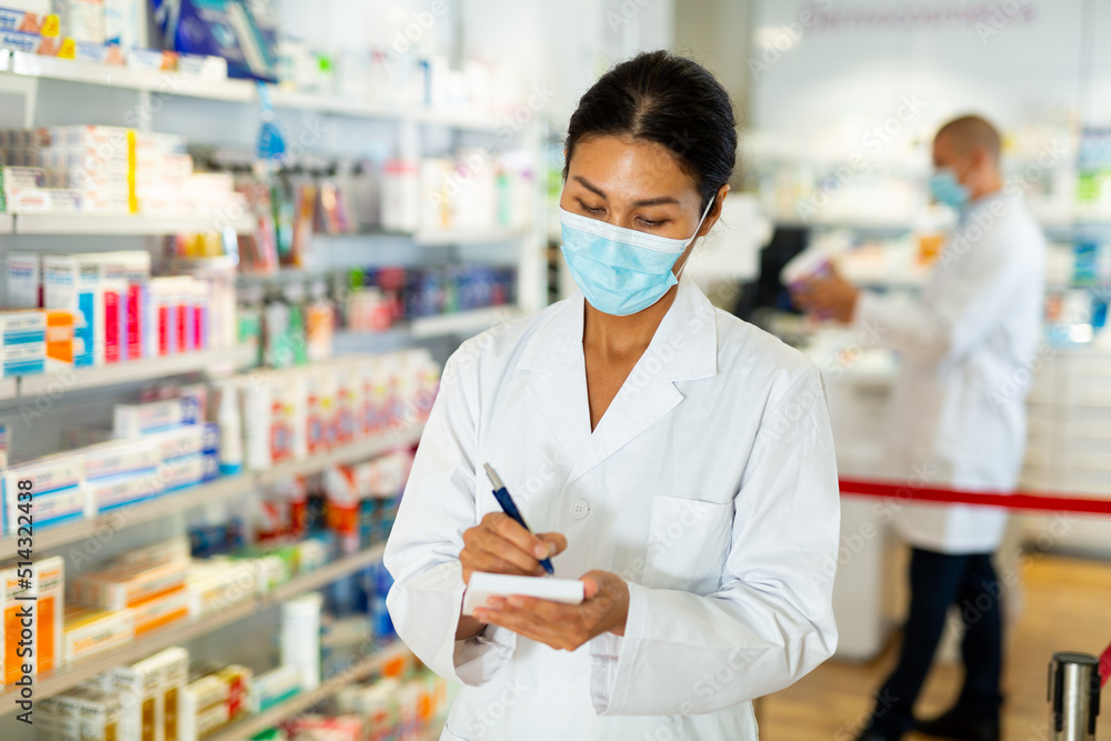 Portrait of female pharmacist in protective mask, working in pharmacy during the pandemic, standing in trading floor and makes important notes
