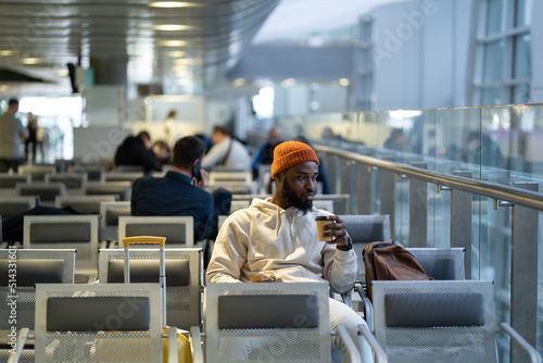 Young African American traveler man drinking coffee and eating sandwich while waiting for flying at airport terminal. Trendy black hipster guy satisfies hunger with a coffee and sandwich. 