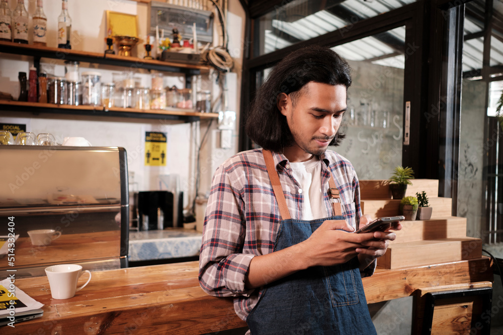 Long hair handsome Asian male startup barista with an apron stands at a casual counter bar cafe, chats about online orders in mobile phone application, and coffee shop service jobs entrepreneur. Long hair handsome Asian male startup barista with an apron stands at a casual counter bar cafe, chats about online orders in mobile phone application, and coffee shop service jobs entrepreneur.