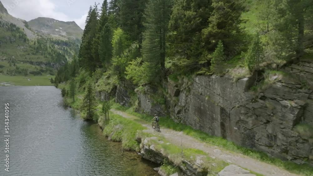 Drone shot of mountain biker on path along lake shore between green mountains in Italy Drone shot of mountain biker on path along lake shore between green mountains in Italy