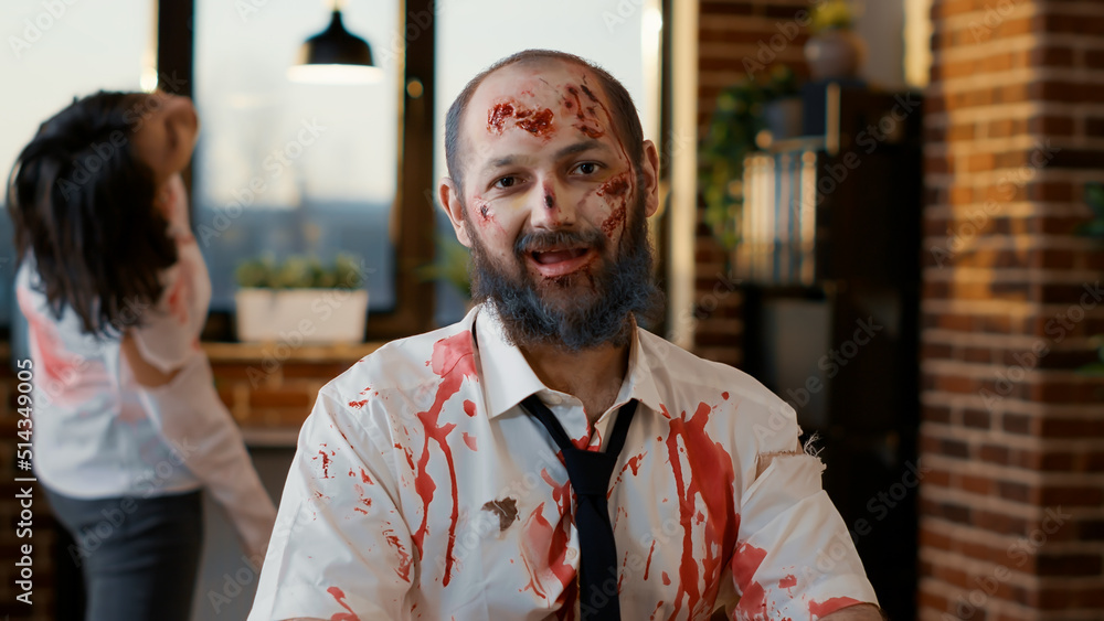 Obraz premium Mindless dumb looking zombie in office workspace sitting at table. Dangerous creepy brain-eating monster with deep and bloody scars and wounds looking at camera while sitting inside company workspace.