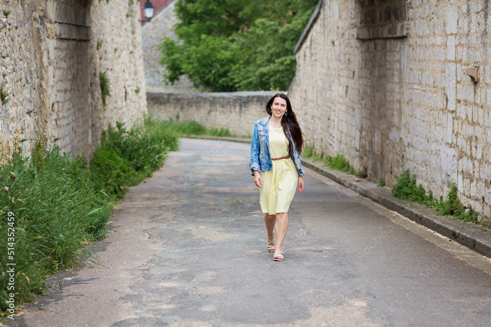 Lifestyle portrait of young stylish woman with long brunette hair walking on the street in old town, wearing yellow dress and denim jacket Lifestyle portrait of young stylish woman with long brunette hair walking on the street in old town, wearing yellow dress and denim jacket
