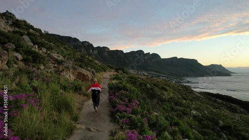 young blonde girl in red jacket running Lions Head Trail at sunset towards 12 Apostles in Cape Town, aerial