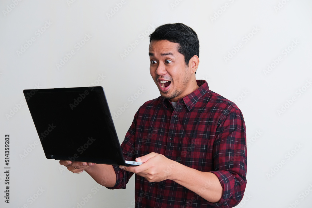 Adult Asian man showing surprised expression when looking to his laptop screen Adult Asian man showing surprised expression when looking to his laptop screen