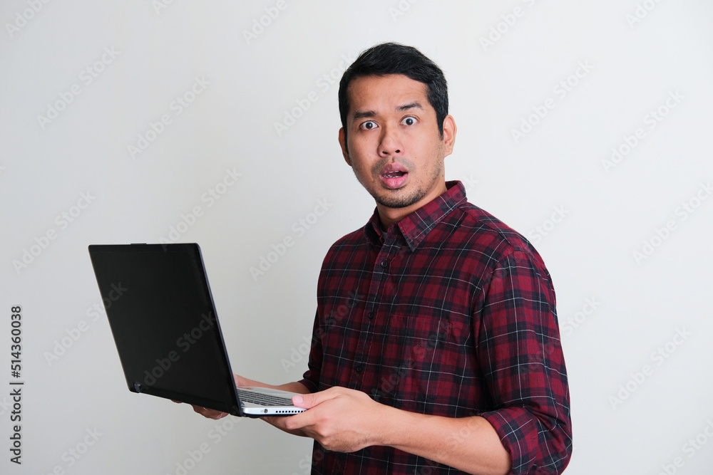 Adult Asian man looking camera with worried expression while holding his laptop Adult Asian man looking camera with worried expression while holding his laptop