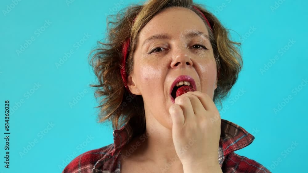 Forty-year-old pin-up woman in a plaid shirt eats strawberries on a blue background. Pin-up woman portrait. Slow motion portrait Forty-year-old pin-up woman in a plaid shirt eats strawberries on a blue background. Pin-up woman portrait. Slow motion portrait