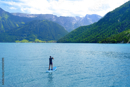man sailing on sapboard, lake Achensee in Austria, green mountains rises above calm expanse of water, concept of vacation by reservoir, resort place tyrol, active lifestyle
