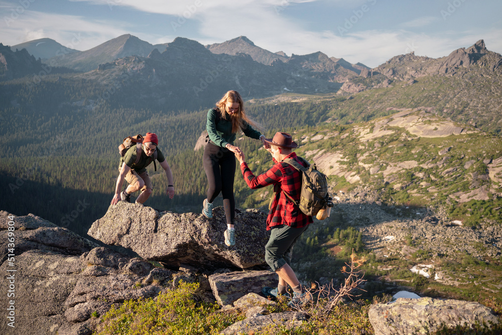 Fototapeta premium Group of young tourists two guys and girl with backpacks in mountains go along route