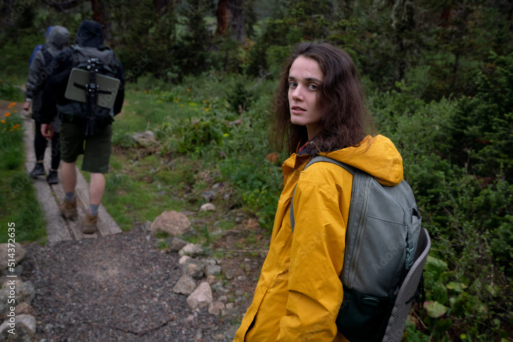 Beautiful young girl in raincoat with backpack looks back on hike in forest Beautiful young girl in raincoat with backpack looks back on hike in forest