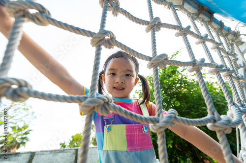 Happy Little Asian child girl playing and climbing rope net at outdoor playground in the park on summer vacation. Kindergarten children kid enjoy and fun outdoor activity and exercising at park.